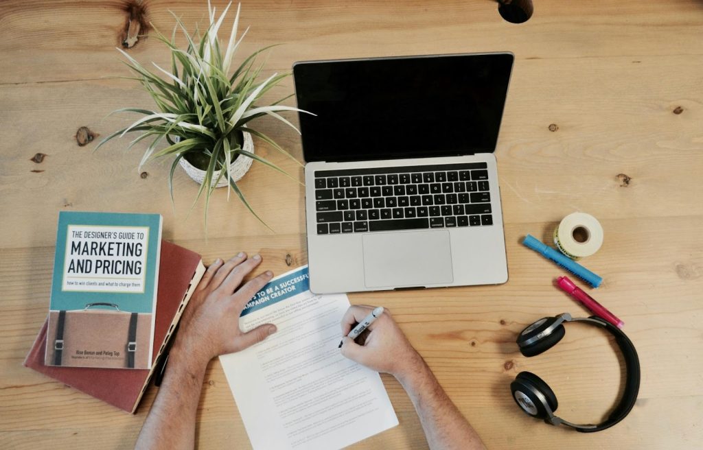 a person writing on a notebook next to a laptop and headphones